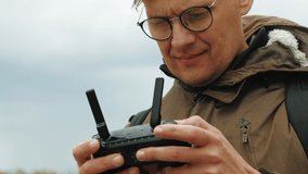 Mature Man wearing glasses in the city on the embankment of the river controls the drone using a remote control, making aerial shooting. Close up portrait. Cloudy weather, spring or cold summer