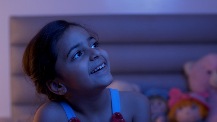 Pretty young Indian child girl watching stars from the window of her room at night. Stock video of a beautiful cute kid sitting in casual wear - looking outside from the window