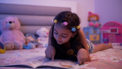 Cute little child girl doing homework and reading a book - Education and smart learning concept. Indian stock footage of a kid enjoying and learning at home. Girl laying on the bed wearing casual