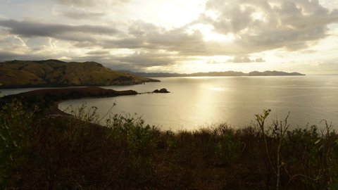 An amazing time-lapse sunset on Maumere Flores beach from the hill with foreground curves of the coastline, mountains and fast moving dark clouds. A scenic golden hour panorama in Indonesia