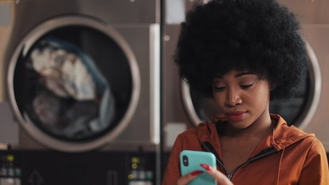 Attractive young African American woman using smartphone sitting in the self-service public laundry. She is wearing a wireless earphones indoors.
