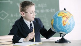 schoolboy in glasses writing in notebook, pointing with finger at globe while sitting near books