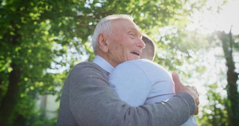 Slow motion of happy grandson is giving an affective hug to his grandfather as a sign of love and respect in a green park on a sunny day.