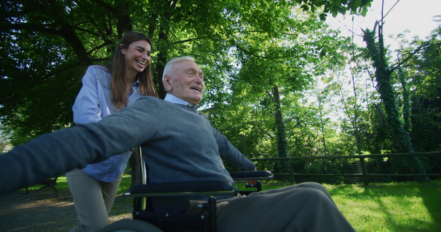 Slow motion of carefree and happy granddaughter and grandfather in a wheelchair having fun to run in a green park on a sunny day. Royalty-Free Stock Footage #1030112861
