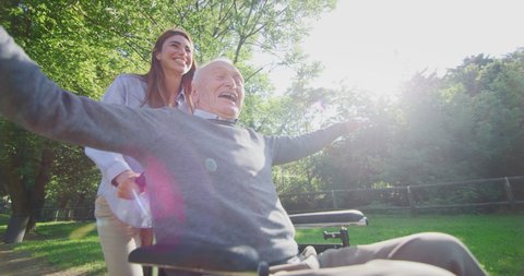 Slow motion of carefree and happy granddaughter and grandfather in a wheelchair having fun to run in a green park on a sunny day.