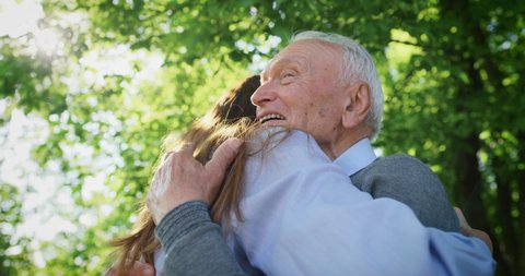 Slow motion of happy granddaughter is giving an effective hug to her grandfather as a sign of love and respect in a green park on a sunny day.