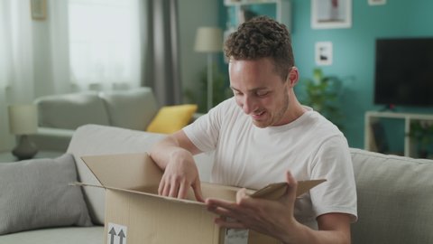 Young man enters his living room with cardboard parcel and open it