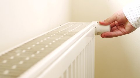 Person's Hand Adjusting Temperature On Thermostat To Control Heat In Central Home Heating System. Radiator near white wall. House in winter concept