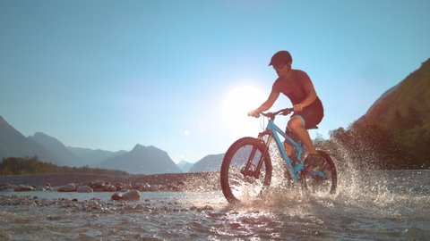 SUPER SLOW MOTION, LOW ANGLE: Fit man rides an e-bike in the shallow river and splashes water around him on a sunny summer day. Active male tourist rides a mountain bike in sunlit Soca river valley.