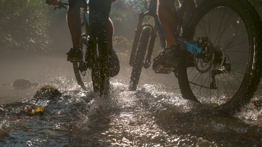 SLOW MOTION, LOW ANGLE CLOSE UP: Two unrecognizable persons riding new mountain bikes across the sunlit stream. Bright sun rays illuminate the river as two men ride bikes through the dark forest.
