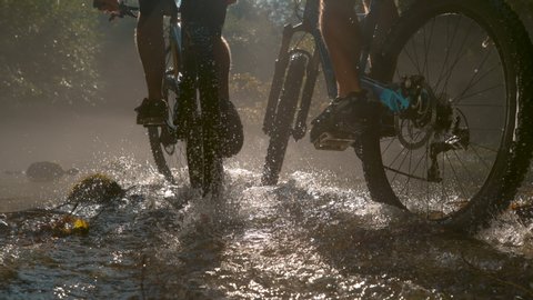SLOW MOTION, LOW ANGLE CLOSE UP: Two unrecognizable persons riding new mountain bikes across the sunlit stream. Bright sun rays illuminate the river as two men ride bikes through the dark forest. : vidéo de stock