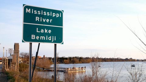 Road Sign at bridge over Mississippi River at Lake Bemidji in early morning Fishing Opener with people trying to catch fish on pier and in boats and town of Bemidji Minnesota in background.