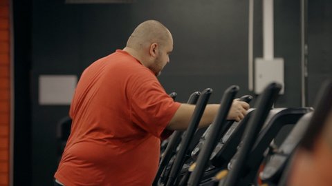 Tired and overweight man wearing in orange tee-shirt clothes standing inside modern and large sport gym. He walking on professional orbitrek for cardio intensity and endurance improvement