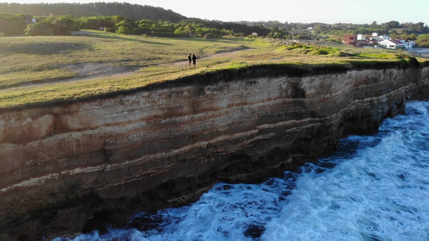 Aerial static 4k drone shot of two guys standing on the edge of a cliff, while waves are breaking in, on a sunny afternoon with a clear blue sky background inMar del Plata, Argentina