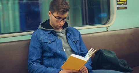 Nice guy with headphones and wearing glasses reads a book in a subway car. Portrait view.