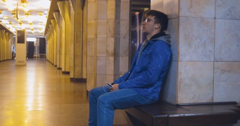 Guy sits on the bench of the subway station and listens to music on headphones. Young man with glasses is waiting at the metro station.