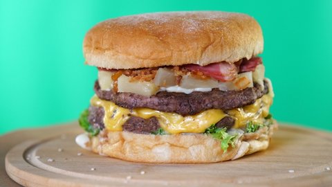 Beef burger with cheese, sauce and vegetables rotating on a wooden board in front of green screen background. Chroma key. Seamless loopable shot