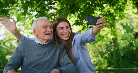 Slow motion of happy granddaughter and grandfather in a wheelchair are having fun to take a selfie photo with cellular phone or making the video call in a green park on a sunny day.