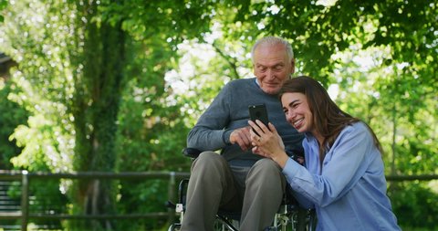 Slow motion of  happy granddaughter and grandfather in a wheelchair are having fun to look their photos or navigating in internet in cellular phone in a green park on a sunny day.