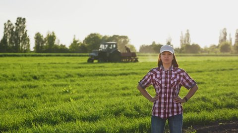 Portrait of a smiling girl farmer in a white cap in a field in spring against the background of a tractor driving across the field.