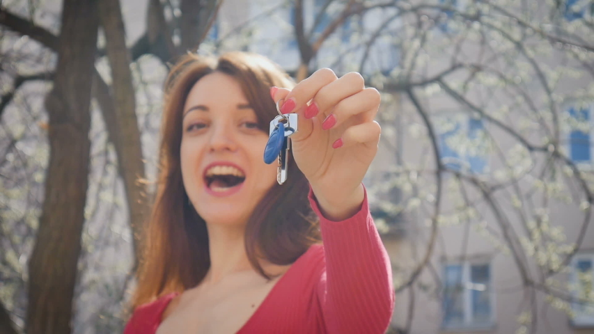 Emotional cazy young woman holding keys with blue keychain with happiness and excitings. Female hand with red manicure holding key outdoors during spring time on urban background.