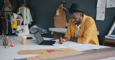 Male fashion designer African American guy is drawing new clothes using laptop creating fashionable garment sitting at table in studio. Work and art concept.