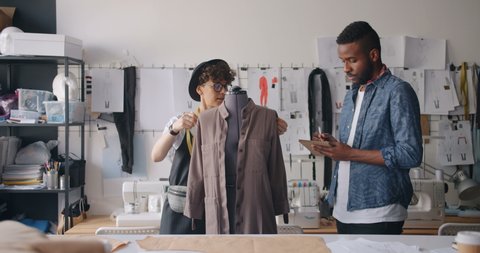 African American man and Caucasian girl tailors are measuring clothes on dummy writing measurements in notebook working in modern studio. People and work concept.
