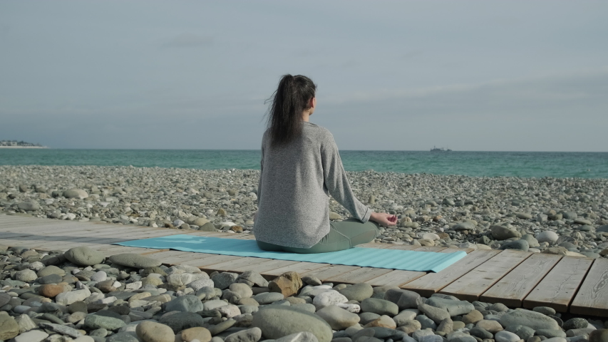 young women wearing sports clothes and sitting in lotus position meditating in the quiet scenery of summer beach. Shot from behind of woman meditating on a mat.