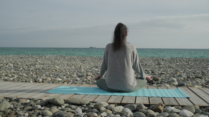 Back view shot of a young woman meditating in lotus pose on pebbl sea beach. Sporty brunette girl trying to find inner peace, yoga in nature.