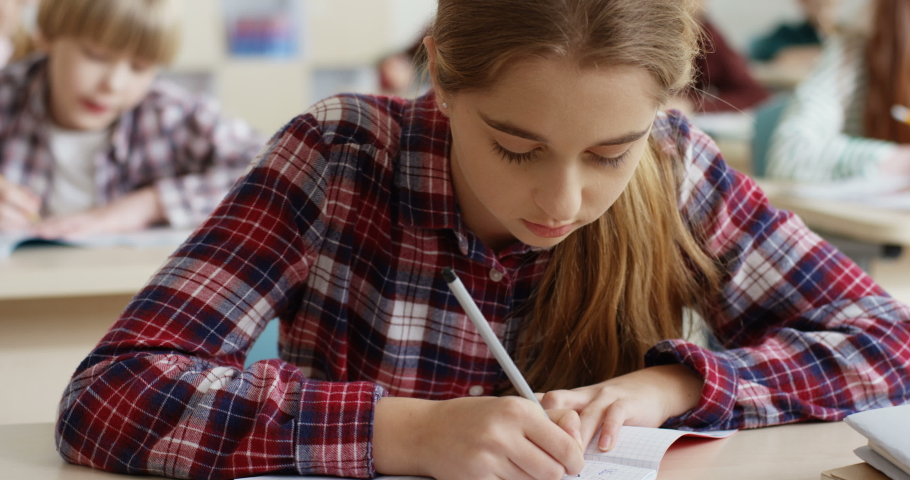 Close up of the Caucasian teen schoolgirl with fair hair writing in the copybook an exercise and thinking at the lesson at the exam.