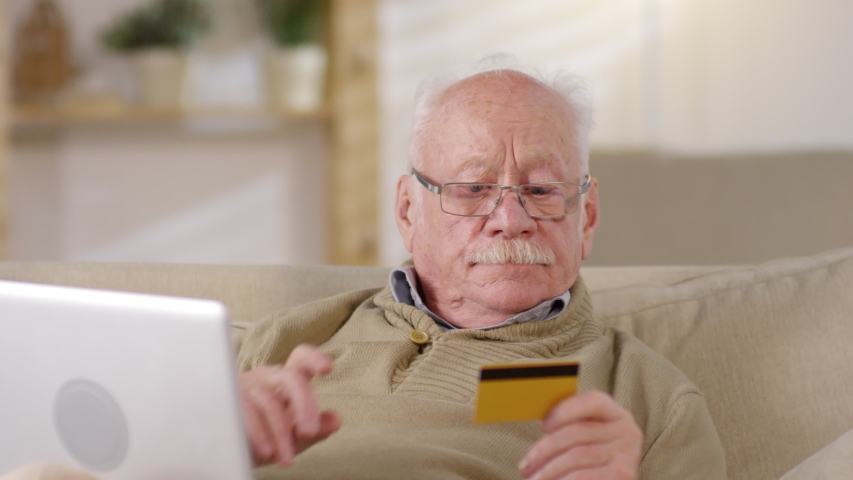 Close up face of concentrated elderly man in glasses making online payment using credit card and laptop computer