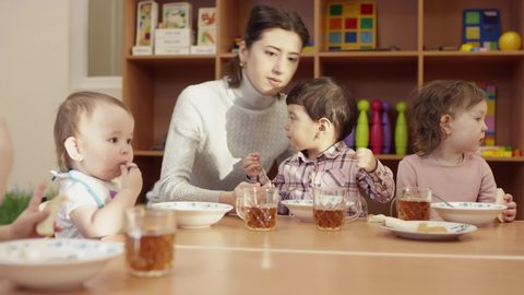 kindergarten teacher sits at the table with the children, and feeds the little dark-haired boy with a spoon, the other children eat on their own, the table is full of plates and mugs, in the