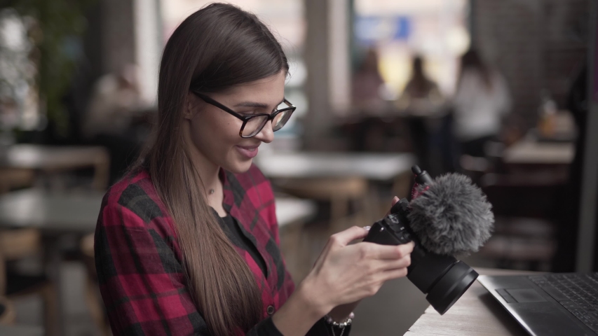 A beautiful female photographer holds her own camera and checks made photos from recent photoshoot. She is very satisfied and smiles. There is grey mini microphone on the camera. Background is blurred