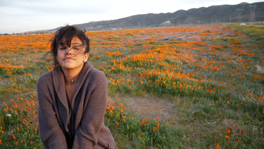 A cute young girl sitting in a field of wild flowers smiling and laughing at golden hour sunset as her hair is blowing in the wind SLOW MOTION.