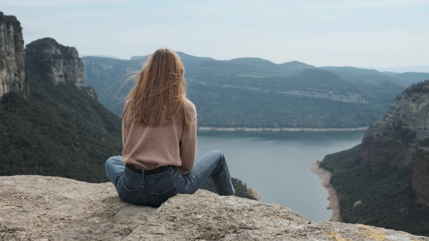 Thoughtful girl in glasses sits on top of a mountain and enjoys a wonderful view of the blue lake and beautiful mountains,  hair weaves wind, happy young girl life