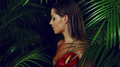Beautiful girl with tanned skin holds  red exotic flower in her hands being among the tropical plants. Natural makeup and wet hair in girl amazon. Natural cosmetics, spa procedures.