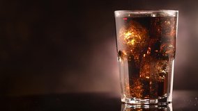 Cola with ice cubes close-up. Cola with Ice and bubbles in glass. Soda closeup. Food background. Rotate glass of Cola fizzy drink over brown background. Cocktail. Slow motion 4K UHD video footage