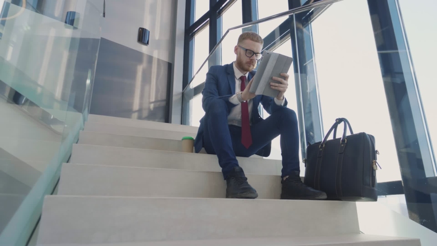 A thoughtful businessman sits on a staircase in lobby with tablet and a cup of coffee | Shutterstock HD Video #1030207112