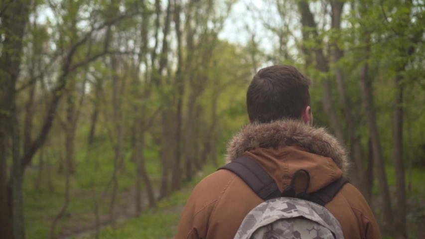 A man in a warm broun jacket walks through the woods. The man goes in front, the camera follows his, then the man makes a circle, looking into the camera. Slow motion video.