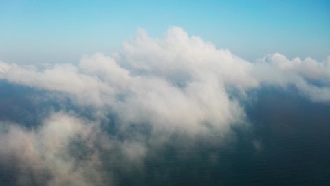 Moving white clouds blue sky on coastline and sea surface scenic aerial view. Drone flies forward high in blue sky through the fluffy clouds on panorama of the sea shore. The sun is hidden. Fog