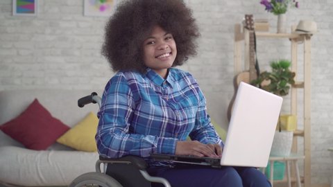 disabled african american woman with an afro hairstyle in a wheelchair uses a laptop looking at the camera