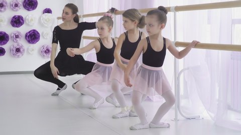 Concentrated girls are learning basic ballet positions in dancing school with careful teacher helping them to master difficult postures.