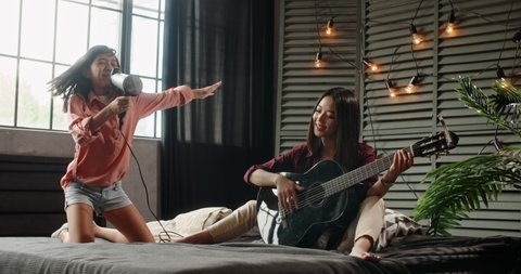 Two asian sisters sitting on bed, elder playing guitar while little kid is singing into hairdryer. Friends having fun spending time together at home - recreational pursuit, family time 4k
