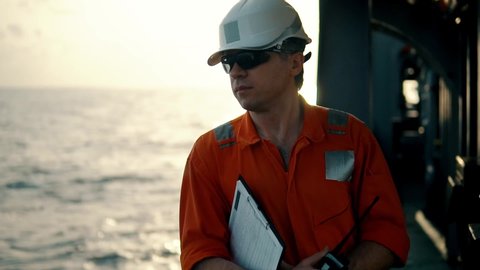 Marine Deck Officer or Chief mate on deck of offshore vessel or ship , wearing PPE personal protective equipment - helmet, coverall. He speaks VHF walkie-talkie radio in hands.