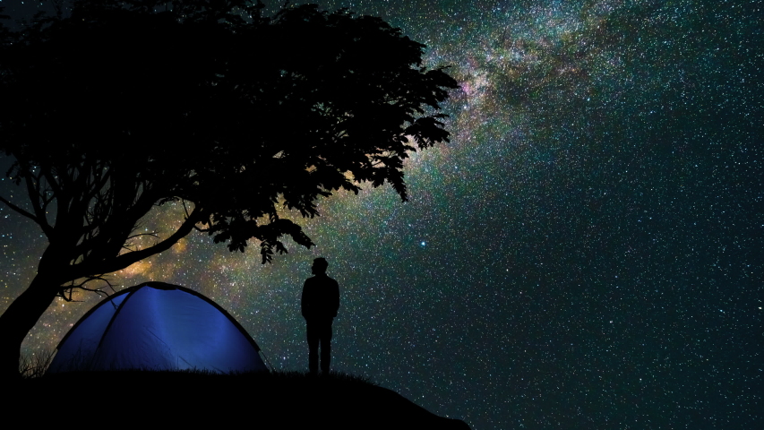 The man standing near a camping tent and tree against the starry sky. time lapse