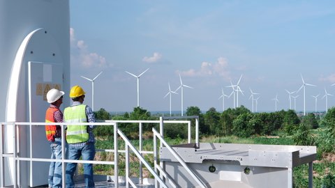 Two Asian engineers are checking the operation of wind turbines in wind farm field for production of electric power