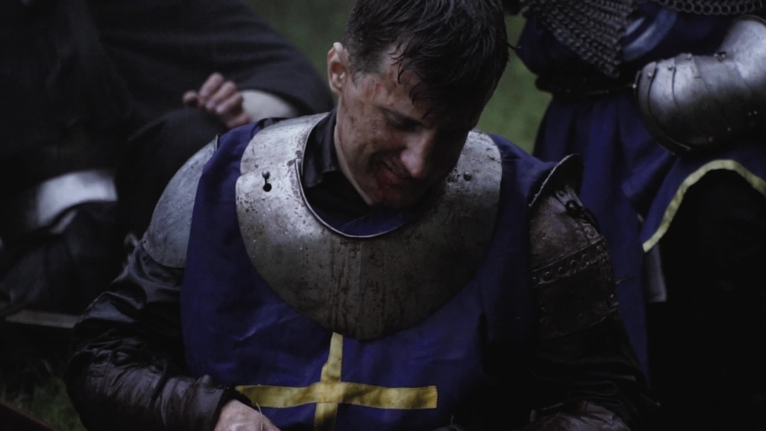 Warrior in blue medieval armor is sitting on the ground and cleaning his sword