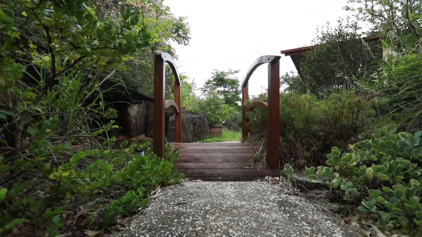 A concrete garden walkway with a wooden bridge, pond, and lawn near a house