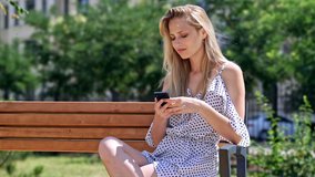 Pretty pleased blondy woman in dress using smartphone while sitting on bench at park