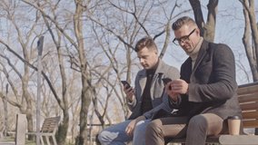 Two smiling adult men speaking using smartphones while sitting in the park
